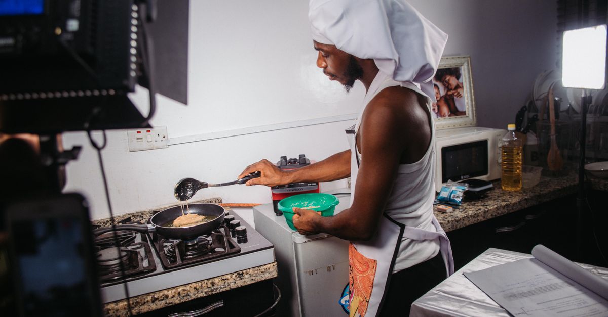 Man in apron and chef hat cooks in home kitchen with professional lighting setup.